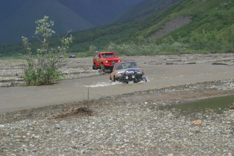 Silver SUV and red truck driving through river in a rocky, mountainous landscape