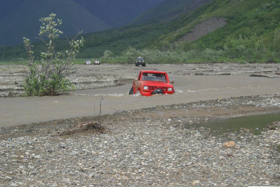 Red pickup truck driving through a shallow river with other vehicles in the background