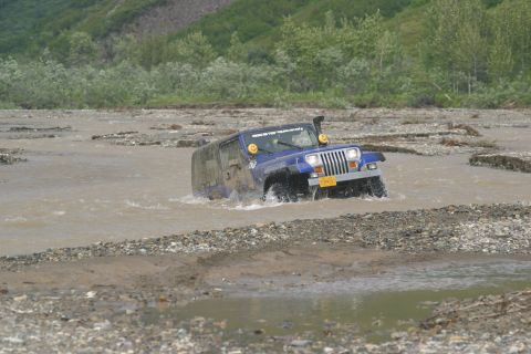 2003 Jeep TJ - A blue Jeep Wrangler navigates a shallow, rocky river, kicking up water as it goes.