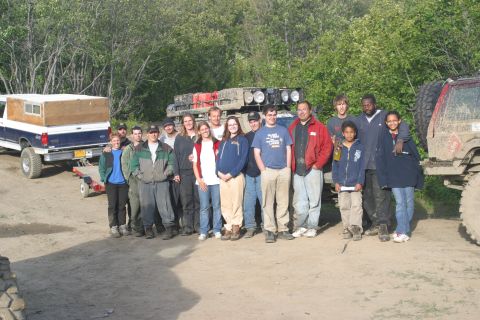 2003 Jeep TJ - A group poses for a photo with their off-road vehicles in a dirt lot surrounded by trees.
