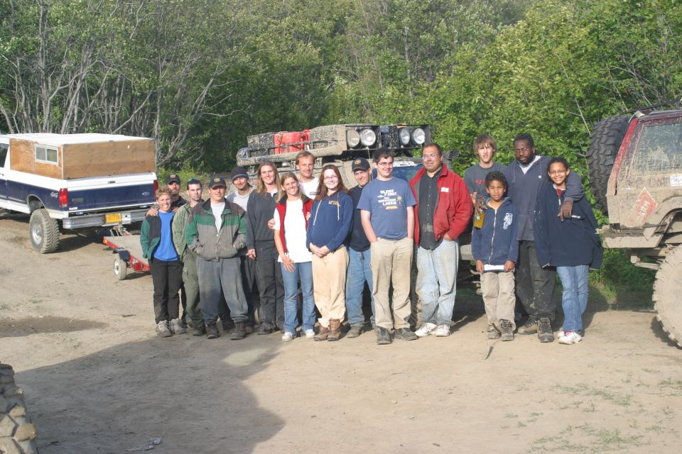 Group of people standing in front of a blue pickup truck with a camper shell, a modified SUV with a roof rack, and a red Jeep-like vehicle