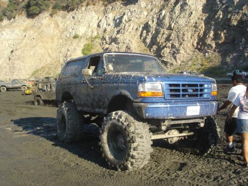 Blue 1991 Ford Bronco, lifted with big tires, covered in mud. Other vehicles and people in background
