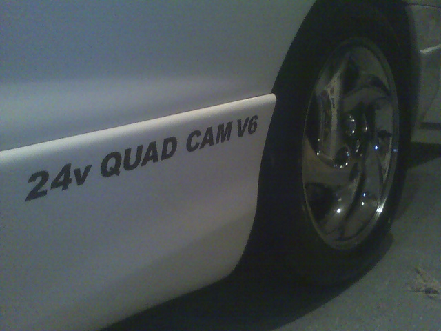 White car side with '24v QUAD CAM V6' decal and chrome wheel