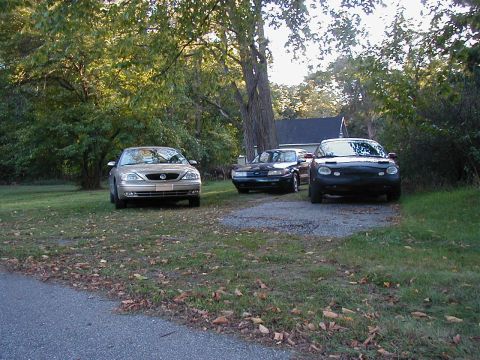 1990 Ford Taurus - Three vehicles are parked on a gravel driveway surrounded by grass and trees.