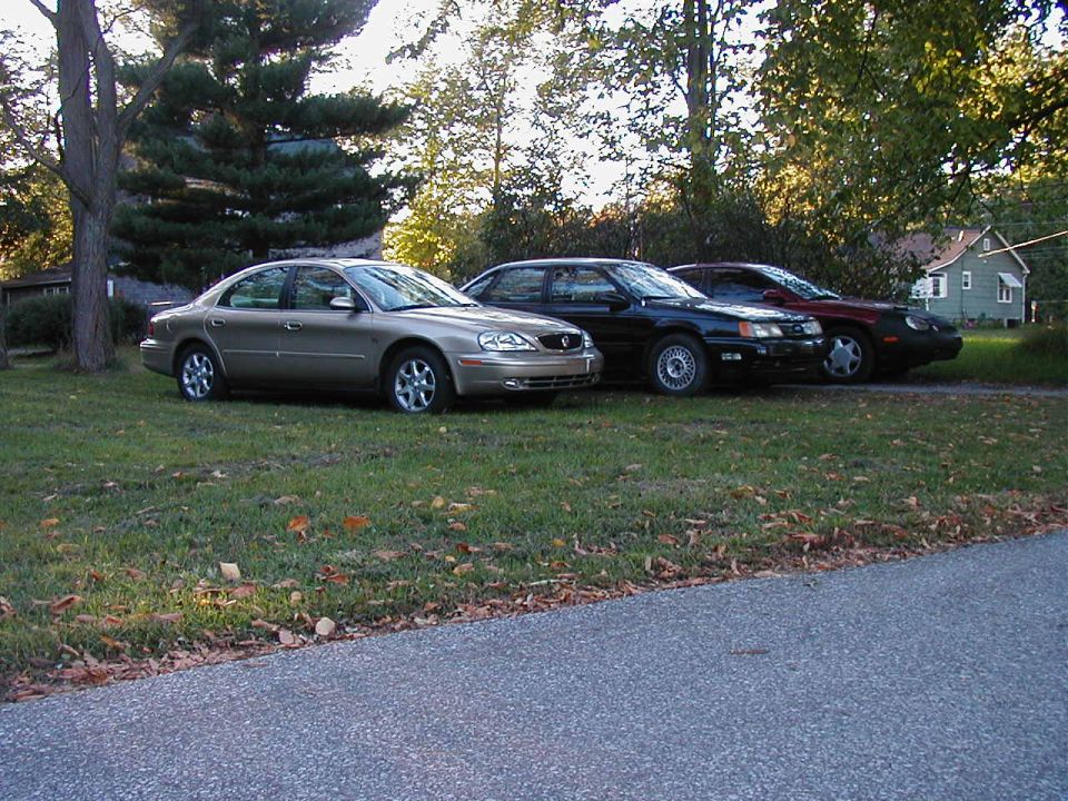 Three sedans parked on a grassy area next to a road, with trees and a house in the background