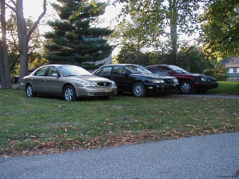 1990 Ford Taurus - Three sedans are parked on the lawn in front of some trees and a house.