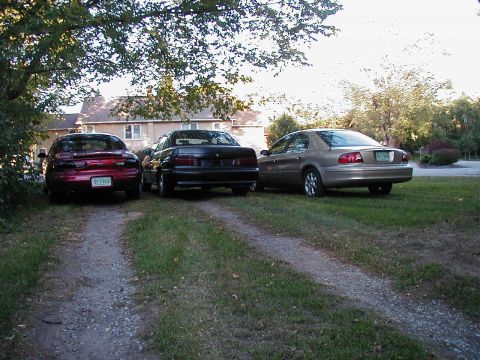 1990 Ford Taurus - Three sedans are parked on a gravel driveway in front of a house, with the gold sedan on the right showing its rear.