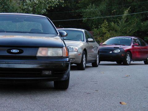 1990 Ford Taurus - Three cars are parked on asphalt with trees in the background. The front car is a black Ford Taurus.