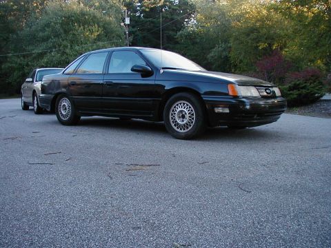 1990 Ford Taurus - A black 1990 Ford Taurus sedan is parked on asphalt, with another car visible behind it.