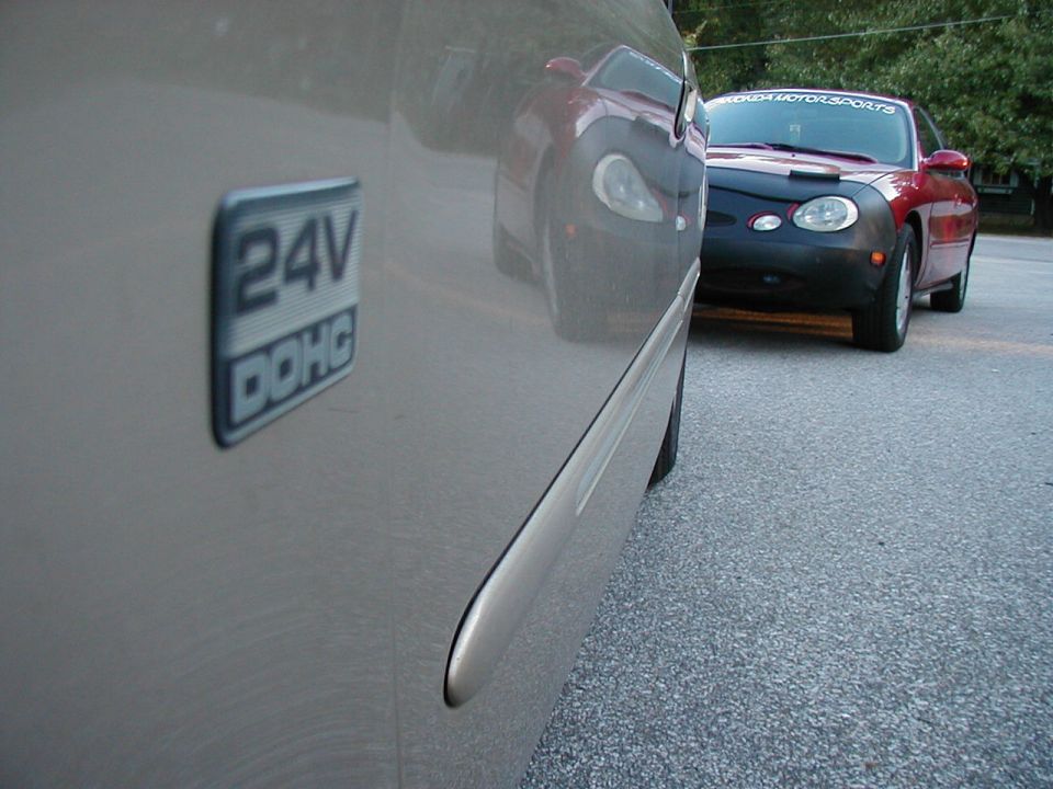 Close-up of a 24V DOHC badge on a car door, with another car visible in the background