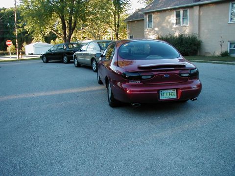 1990 Ford Taurus - A red Ford Taurus is parked behind other vehicles on a residential street.