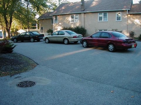 1990 Ford Taurus - Three sedans are parked in a driveway in front of a house, with the closest vehicle being a maroon model with a rear spoiler.