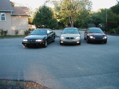 1990 Ford Taurus - Three sedans are parked in a residential driveway. The vehicle on the left appears to be a 1990 Ford Taurus.