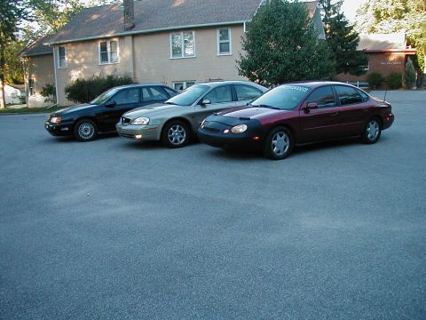 1990 Ford Taurus - Three sedans are parked in a lot, with the closest one being a maroon model with a car bra.