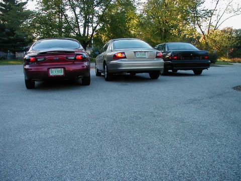 1990 Ford Taurus - Three sedans, including a red 1990 Ford Taurus, are parked in a lot.