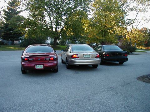 1990 Ford Taurus - Three sedans are parked in an outdoor lot, with trees in the background.