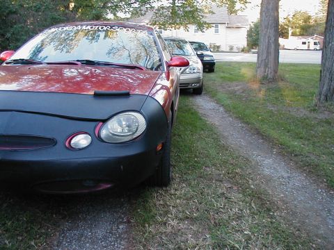 1990 Ford Taurus - A red car with a car cover is parked on a gravel driveway, with two other vehicles behind it.