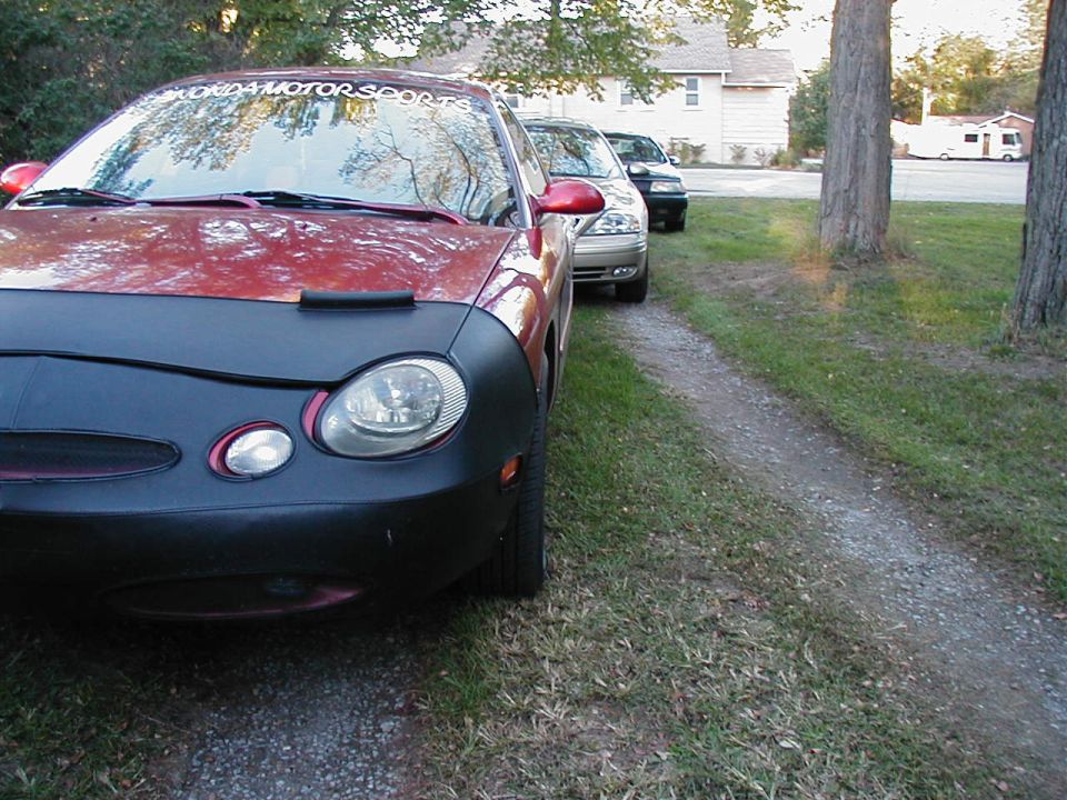 Red car with a black car cover on the front, parked on a gravel driveway. Two other cars are behind it