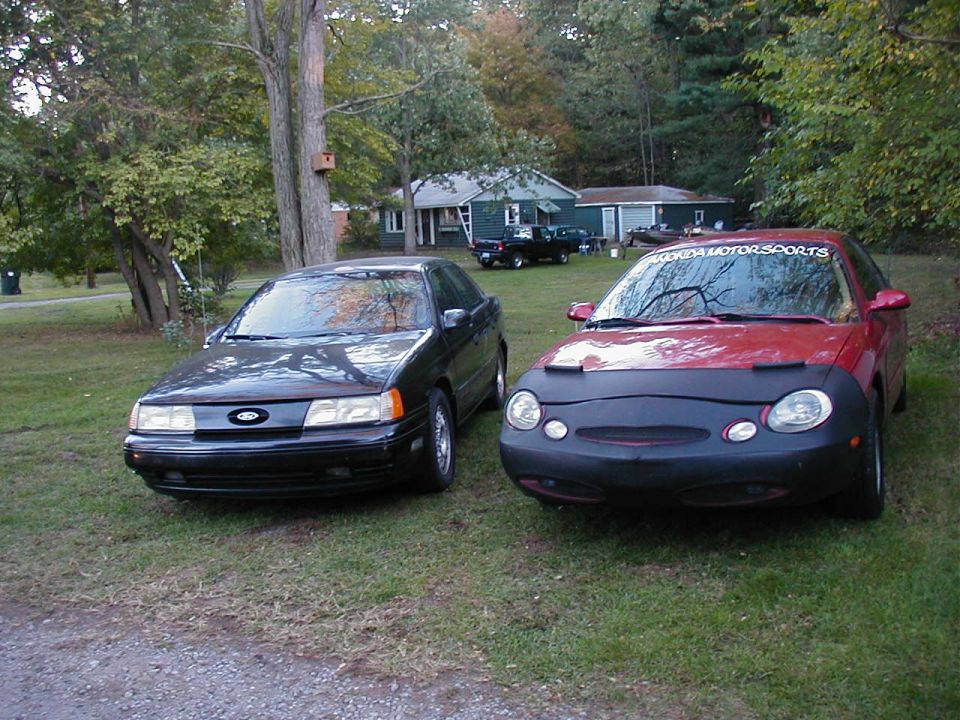 Black and red Ford Taurus sedans parked on grass. Red one has a car bra and a 'Florida Motorsports' banner on windshield
