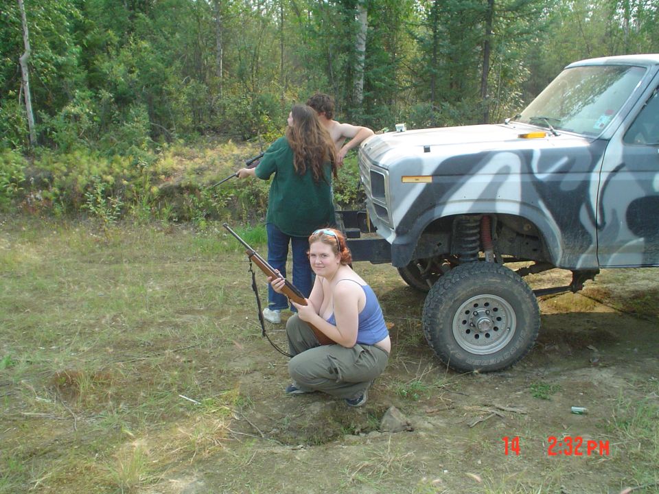 Lifted pickup truck with camouflage paint, two people holding rifles, one crouching