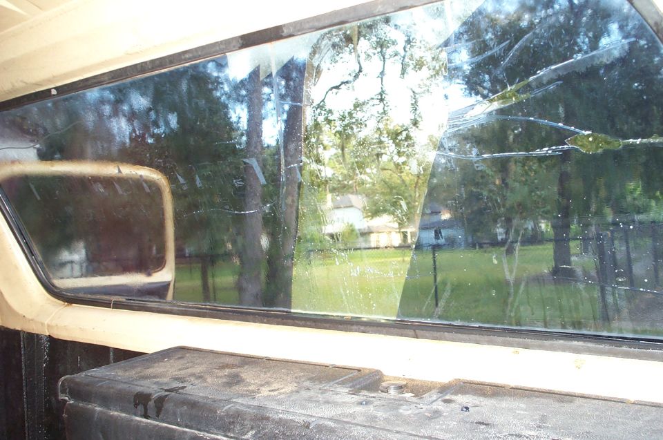Interior view through cracked rear window of a truck, showing trees and a house outside