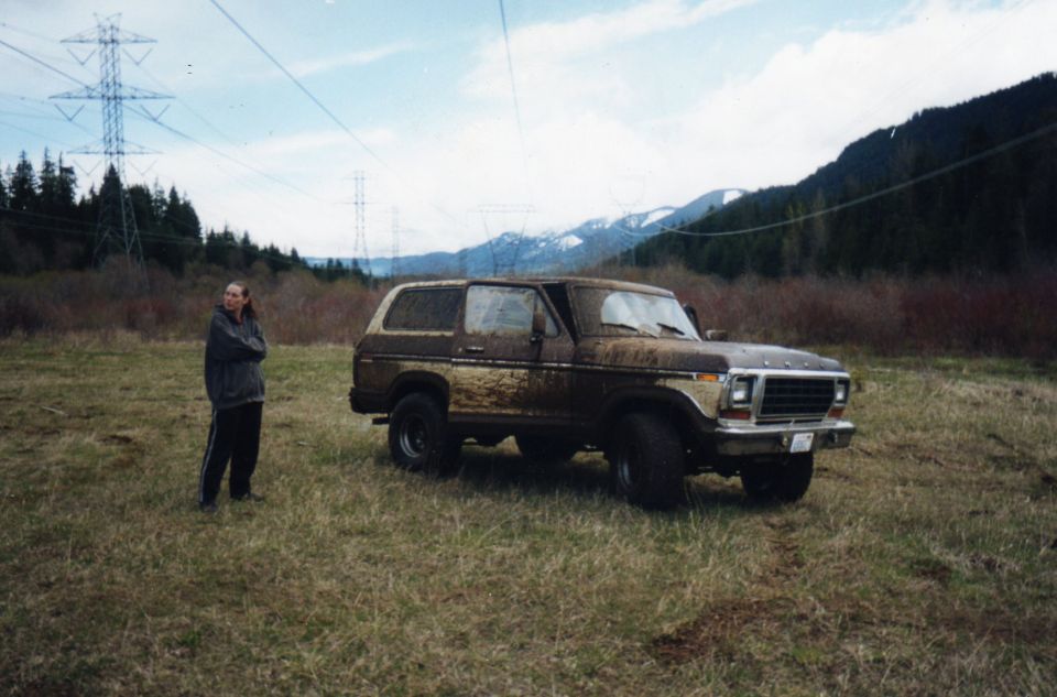 Mud-covered tan Ford Bronco parked in mountain field with person standing alongside