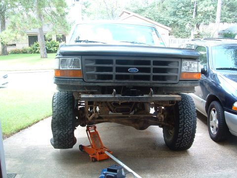 1993 Ford Bronco - The front of a lifted Ford truck is supported by an orange floor jack, with a dark SUV visible to the right.