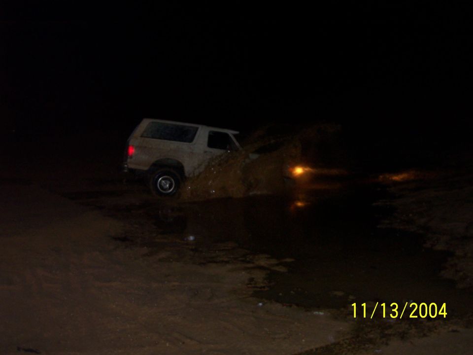 White 1978 Ford Bronco driving through mud at night, splashing water