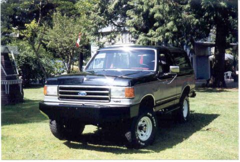 1989 Ford Bronco - This two-tone Ford Bronco is parked outdoors on a sunny day with trees and a building in the background.