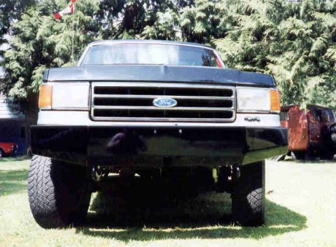 1989 Ford Bronco - Frontal view of a dark truck, a chrome grille with a Ford emblem. Another truck is partially visible in the background.