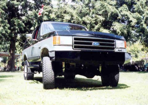 1989 Ford Bronco - A two-tone black-over-silver Ford Bronco/full-size truck shown from a low front three-quarter angle, lifted on large off-road tires and par…