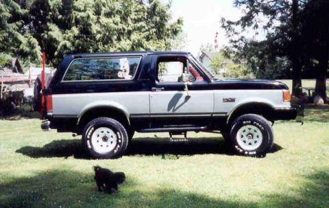 1989 Ford Bronco - Side profile of a two-tone dark blue over silver Ford Bronco full-size SUV with a removable hardtop, white spoke wheels and BF Goodrich all…