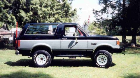 1989 Ford Bronco - A two-tone black-over-silver 1989 Ford Bronco full-size SUV shown in side profile on a grassy lawn, fitted with a suspension lift, white sp…
