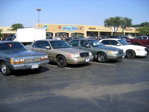 2000 Ford Crown Victoria - Four Ford Panther-platform sedans parked nose-out in a strip-mall lot in front of a "Terra Toys" storefront: a boxy silver-blue 1980s LTD C…
