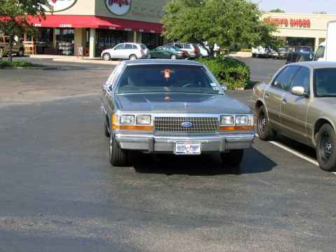 2000 Ford Crown Victoria - A silver sedan is parked in a lot next to another car, with a building in the background.