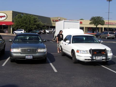 2000 Ford Crown Victoria - Two Ford Crown Victoria sedans parked nose-out side by side in a strip-mall lot, a green-gray car on the left and a white ex-police unit on…