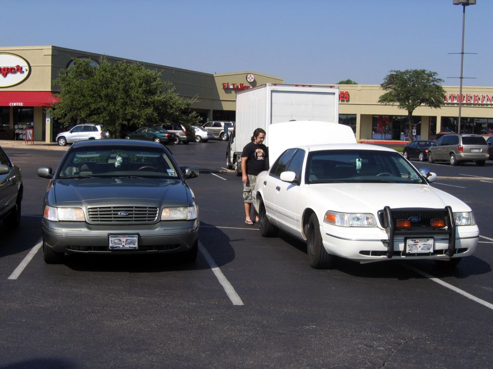Green-gray and white Ford Crown Victoria sedans parked side by side in a strip mall lot