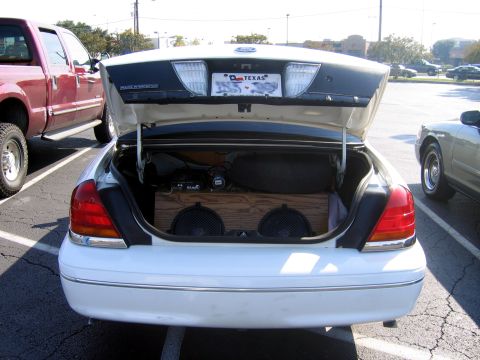 2000 Ford Crown Victoria - The trunk of a white sedan is open, revealing a large wooden box with two speakers inside, alongside other audio equipment.