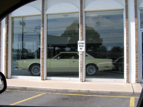 2000 Ford Crown Victoria - A light yellow 1977 Ford Thunderbird coupe is visible through a showroom window, with a 'Customer Parking Only' sign on a pillar.