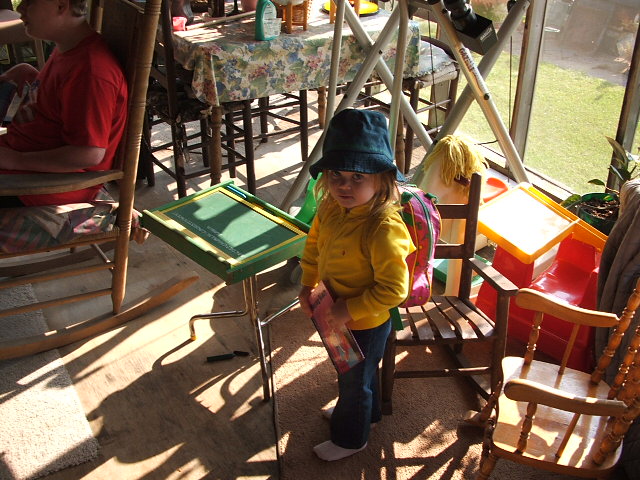 Child in yellow shirt and blue hat with backpack stands in a sunlit room with chairs and a table
