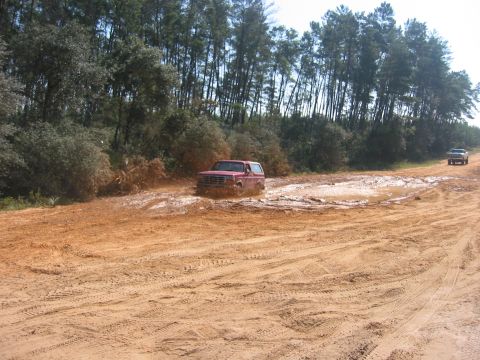 1989 Ford Bronco - A red SUV navigates a muddy track, while another vehicle follows behind on the dirt road.