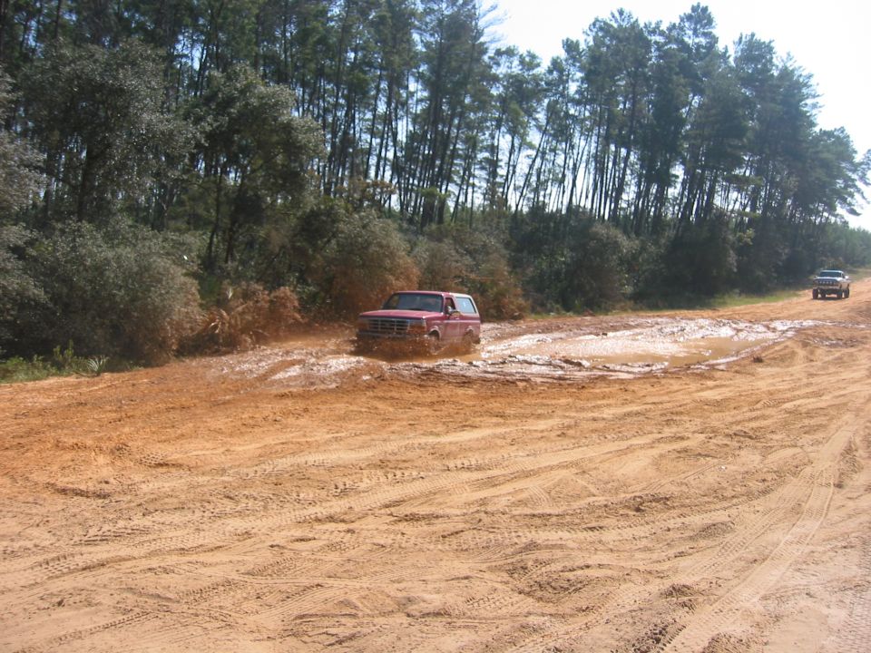 Red SUV stuck in a mud puddle on a dirt road, with trees in the background and another SUV in the distance