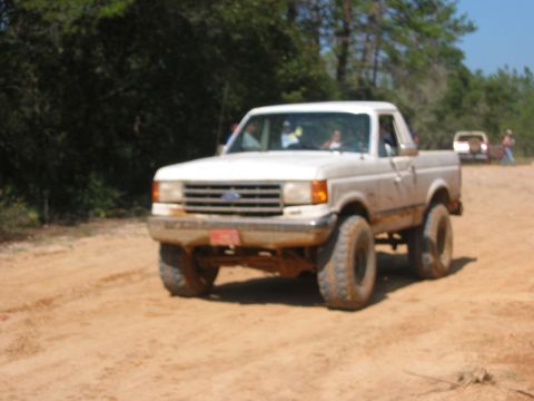 1989 Ford Bronco - A white pickup truck drives on a dirt road, with another vehicle in the distance.