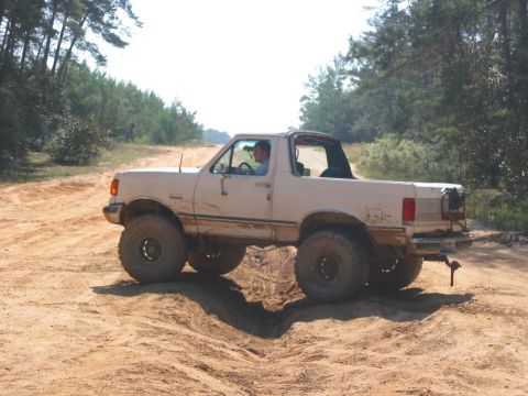 1989 Ford Bronco - A lifted white Ford Bronco navigates a sandy, off-road trail under a clear sky.
