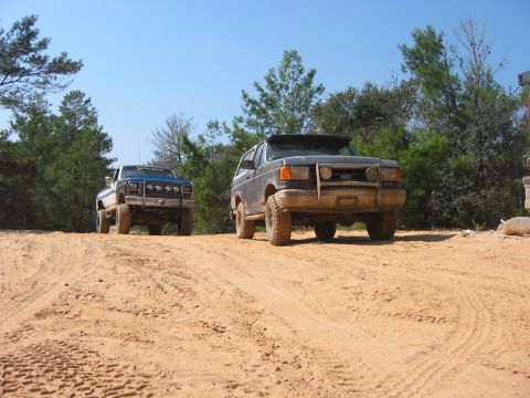 1989 Ford Bronco - Two SUVs navigate a sandy off-road trail under a clear blue sky.