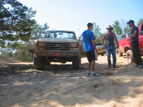 1989 Ford Bronco - Several people gather around a muddy Ford truck in a sandy, wooded area.