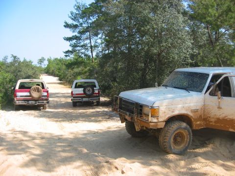 1989 Ford Bronco - Three Ford Bronco SUVs are driving on a dirt road through a wooded area.