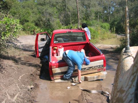 1989 Ford Bronco - A red pickup truck is stuck in the mud with people attempting to free it, while another vehicle is partially visible.