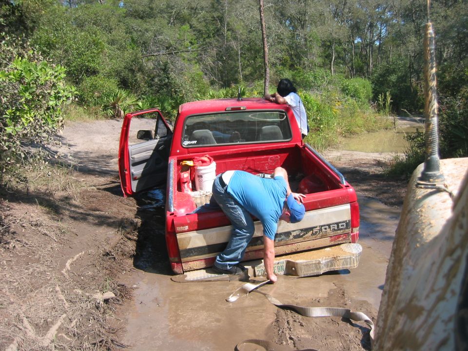 Red pickup truck stuck in mud, driver's door open, people working in the bed. Another vehicle is partially visible on the right