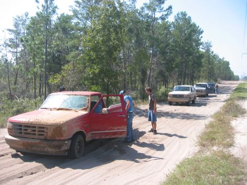 1989 Ford Bronco - A red pickup truck is stuck in the sand on a dirt road, with several other vehicles lined up behind it.